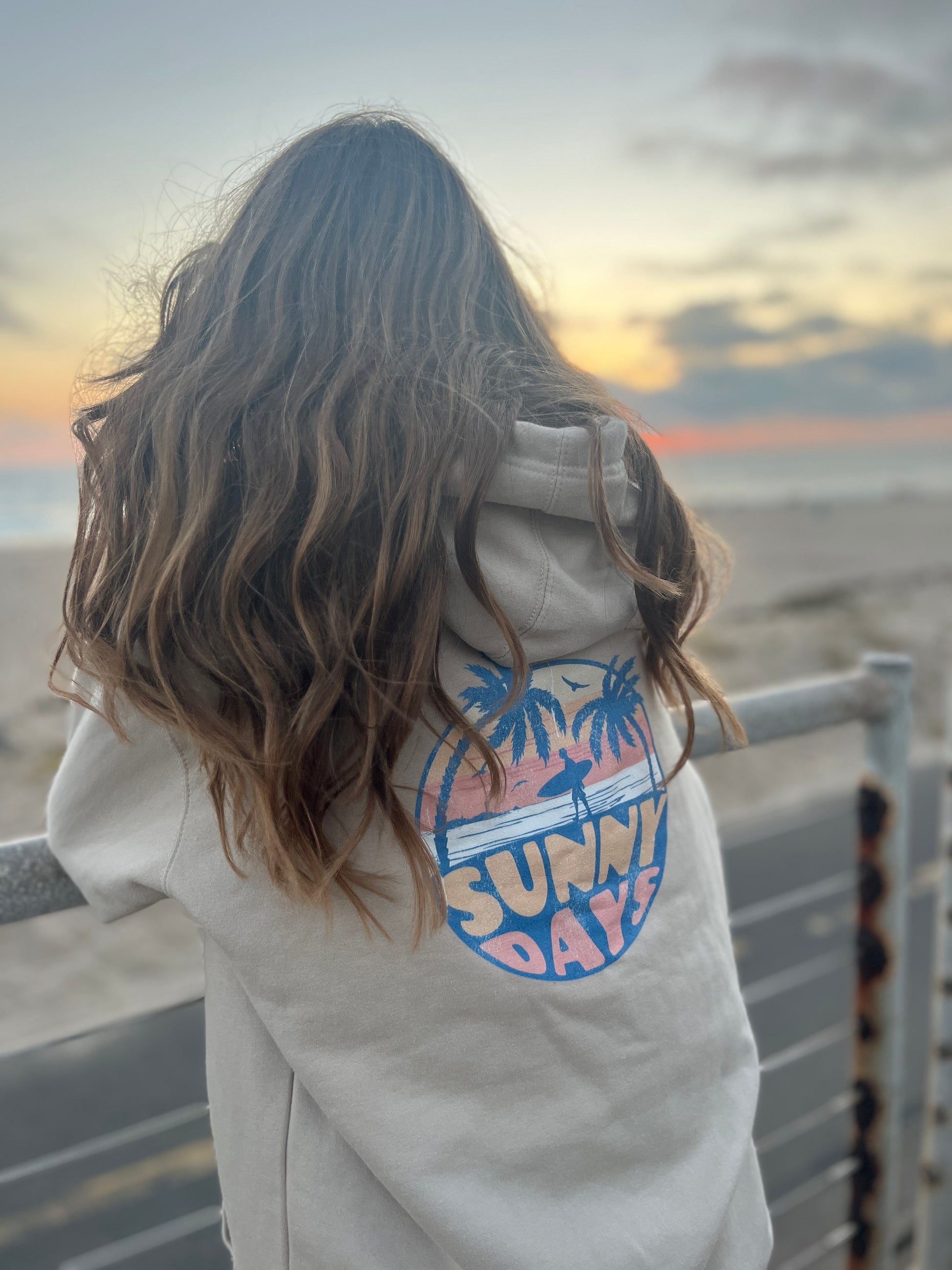 Person wearing a jacket with 'Sunny Days' design at sunset on a beach.