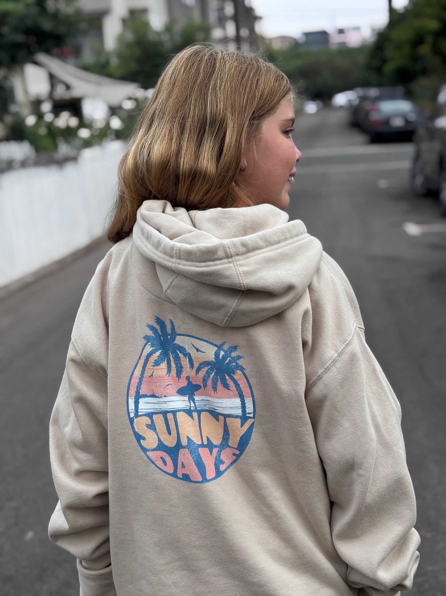 Person wearing a beige hoodie with 'Sunny Days' graphic on the back, standing on a street.