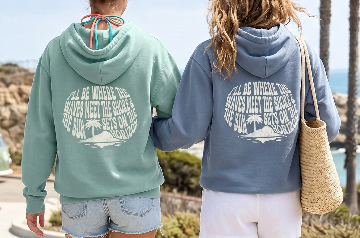 Two people wearing hoodies with a beach-themed design, standing outdoors near palm trees and water.